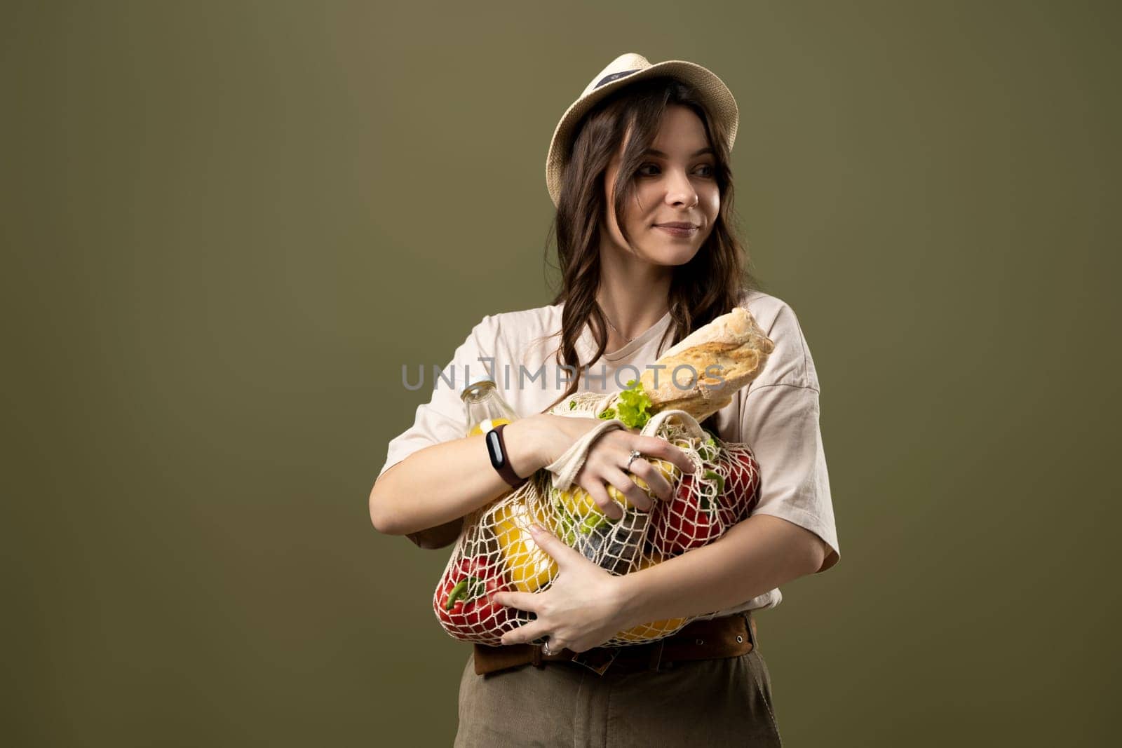 Zero waste, plastic free concept. Brunette woman in beige t-shirt hat holding cotton reusable mesh shopping bags with vegetables, groceries on pastel green background