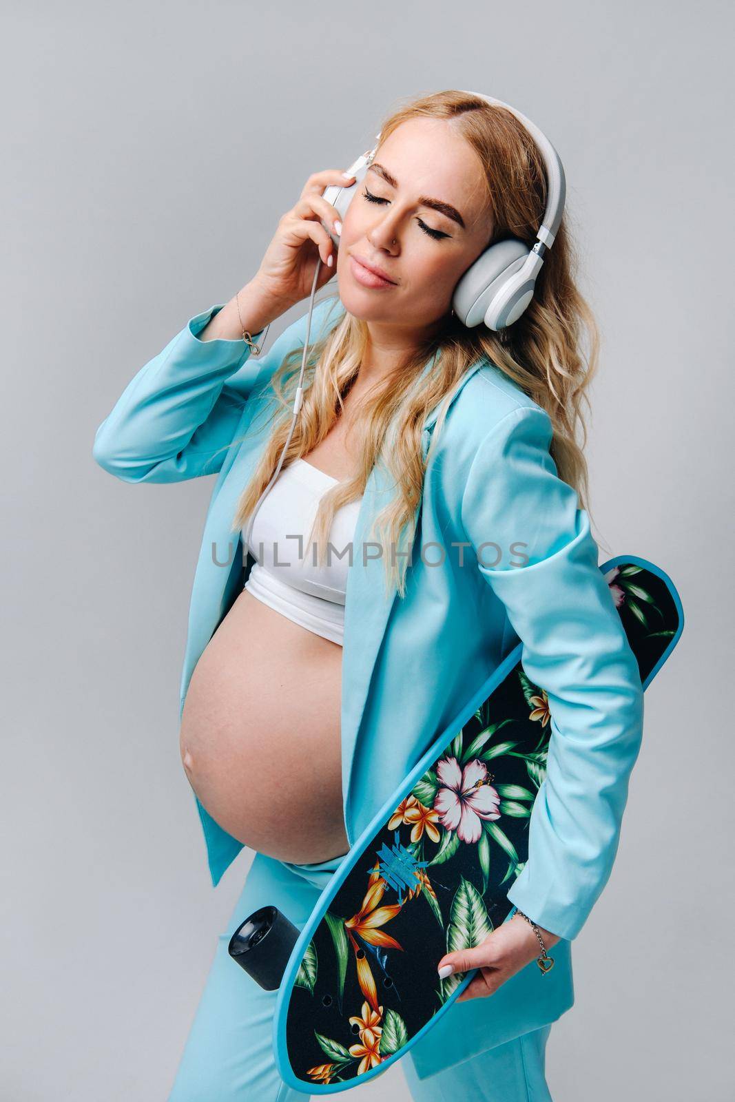 A pregnant girl in a turquoise suit with a skateboard in her hands and headphones stands on a gray background.