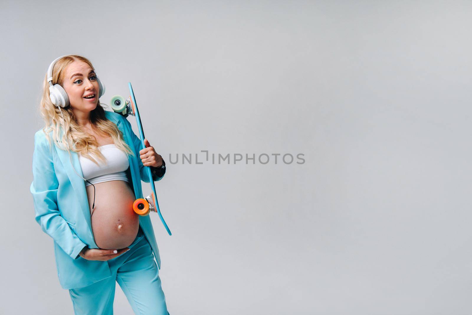A pregnant girl in a turquoise suit with a skateboard in her hands and headphones stands on a gray background.