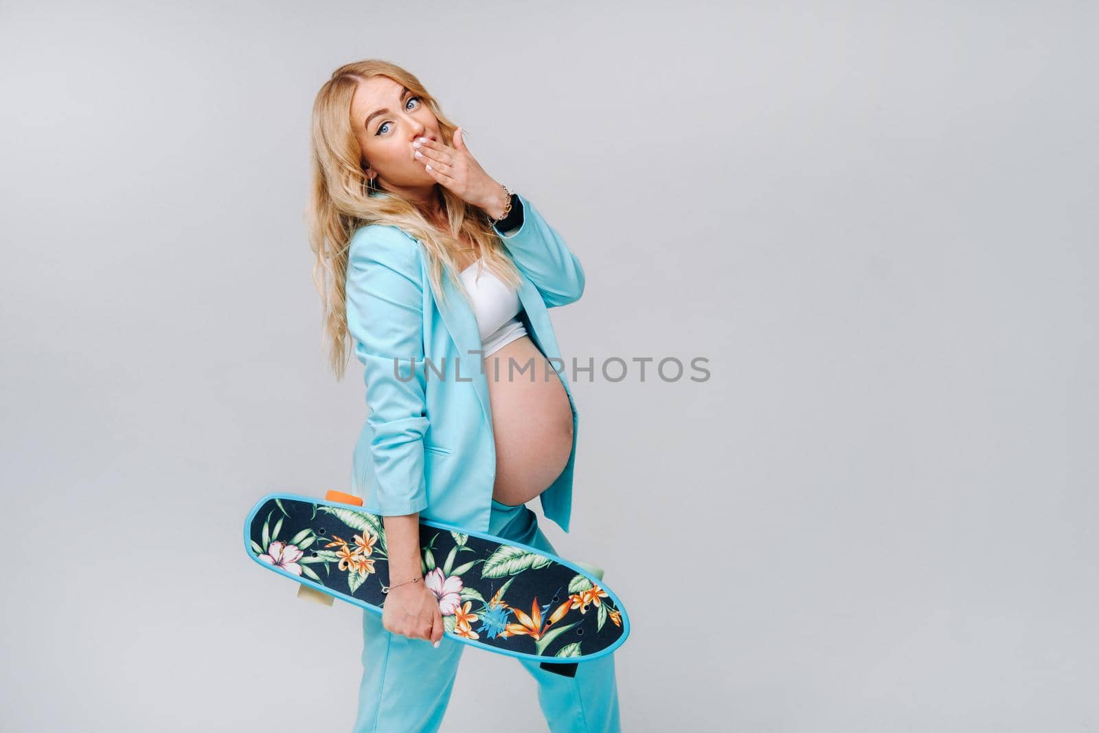 a pregnant girl in turquoise clothes with a skateboard in her hands on a gray background.