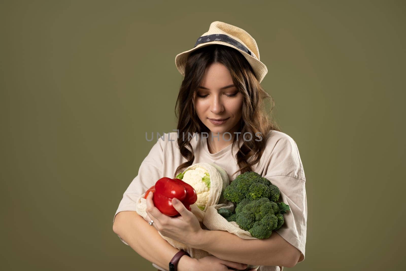 Beautiful brunette woman in beige t-shirt and black hat holding cotton reusable mesh shopping bags with groceries, pepper, broccoli on pastel green background. Zero waste, plastic free concept