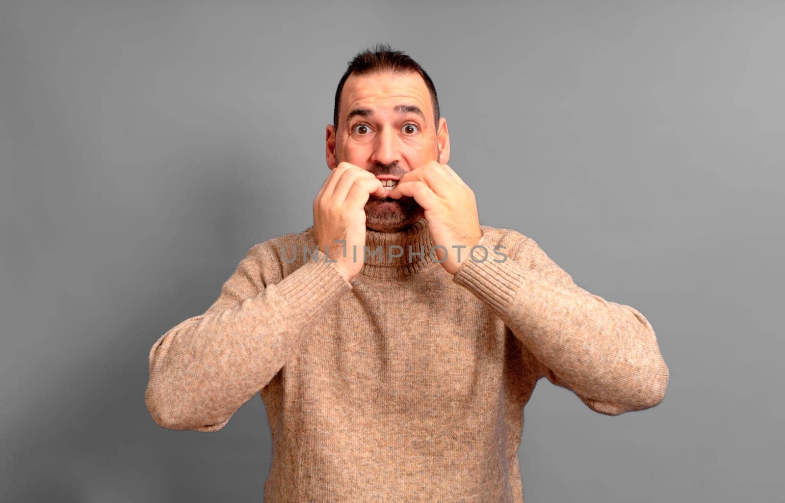 Hispanic man in his 40s scared and shocked biting his nails, staring wide-eyed at something horrible, wearing a turtleneck, standing over gray background, copy space