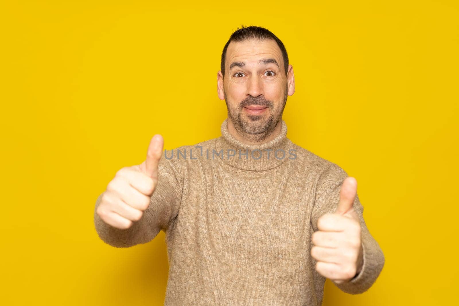 Bearded hispanic man in friendly attitude wearing a beige turtleneck posing funny with thumbs up isolated over yellow background