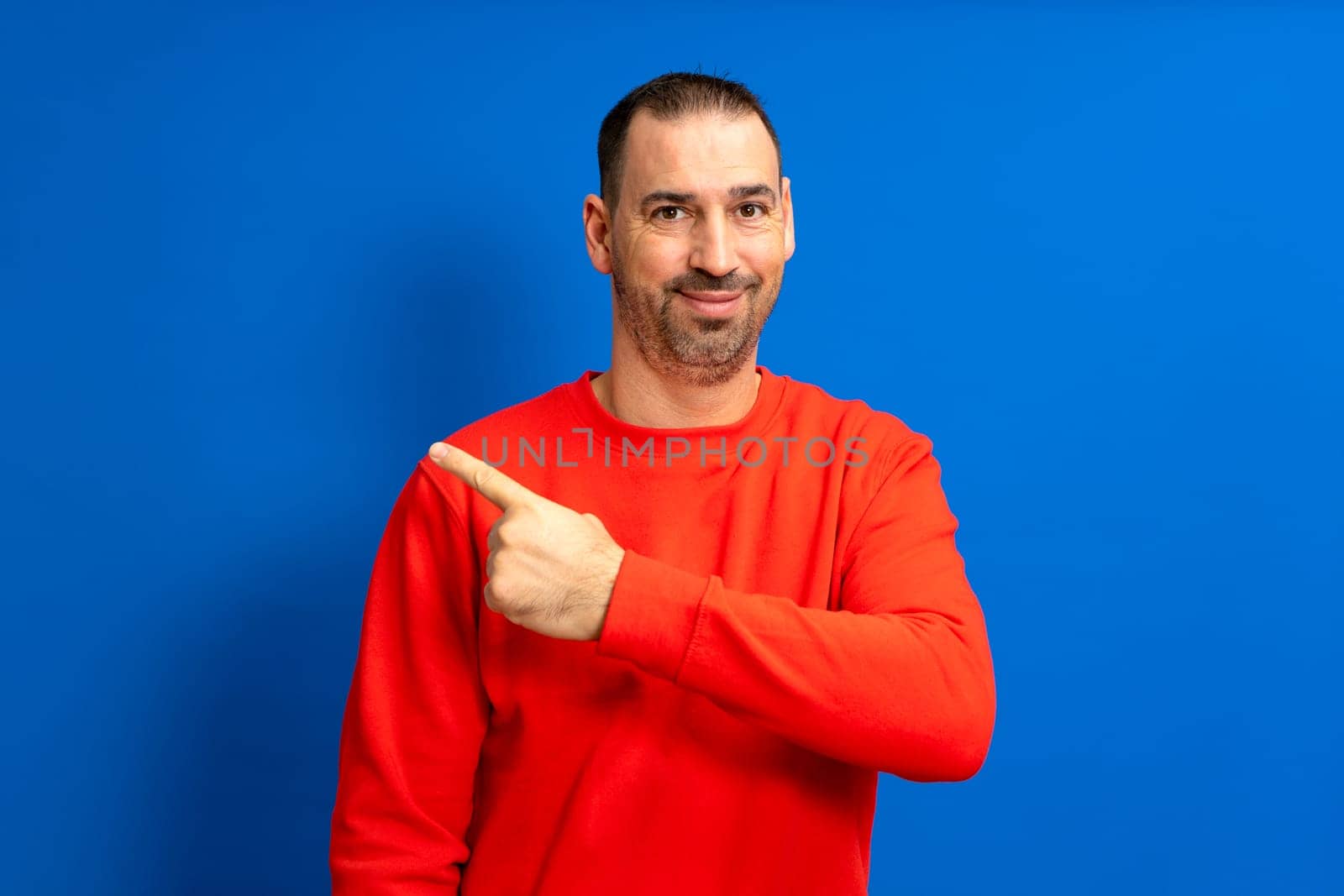 Bearded hispanic man wearing a red sweater smiling at camera while pointing to the side isolated over blue background, indicating a path or direction to follow