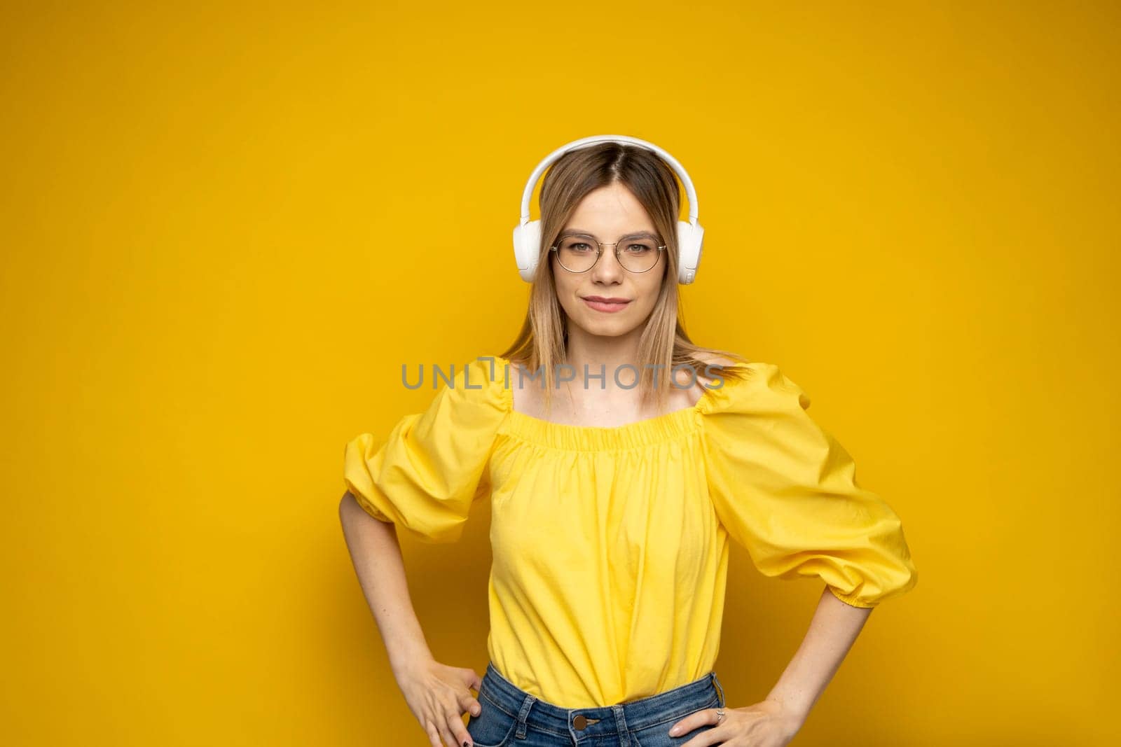 Beautiful attractive young blond woman wearing yellow t-shirt and glasses in white headphones listening music, dancing and laughing on blue background in studio. Relaxing and enjoying. Lifestyle. by vovsht