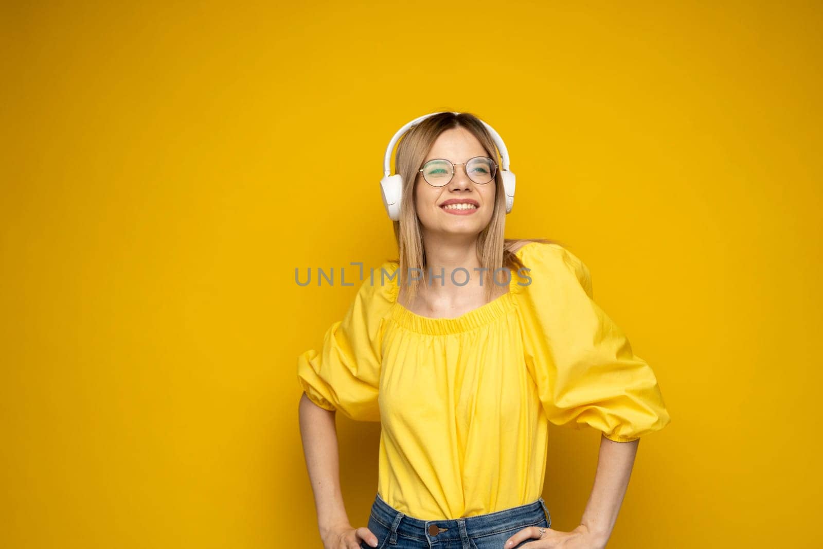 Blonde woman in a glasses and yellow shirt listening music with a headphones. Teenager girl dancing against yellow background