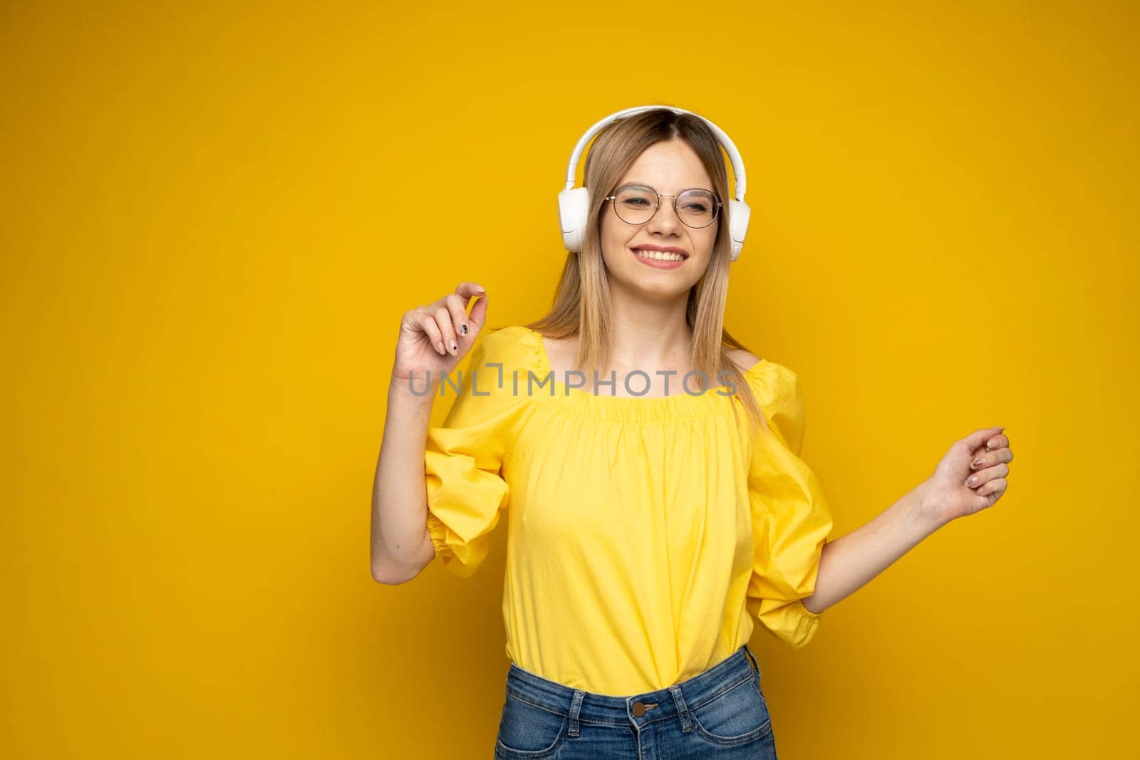 Beautiful attractive young blond woman wearing yellow t-shirt and glasses in white headphones listening music, dancing and laughing on blue background in studio. Relaxing and enjoying. Lifestyle