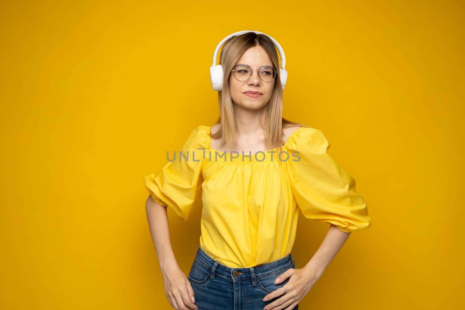 Beautiful attractive young blond woman wearing yellow t-shirt and glasses in white headphones listening music, dancing and laughing on blue background in studio. Relaxing and enjoying. Lifestyle. by vovsht