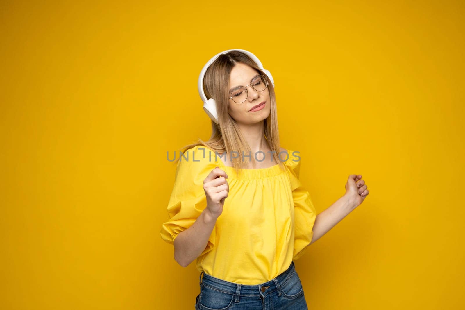 Portrait of charming blonde woman in a yellow t-shirt and glasses wearing white wireless headphones and listening to music isolated on yellow background. by vovsht