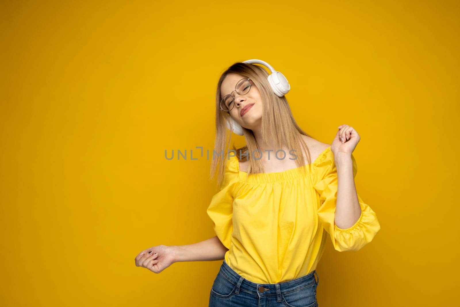 Happy girl in a glasses and yellow shirt dancing and listening to the music isolated on a yellow background