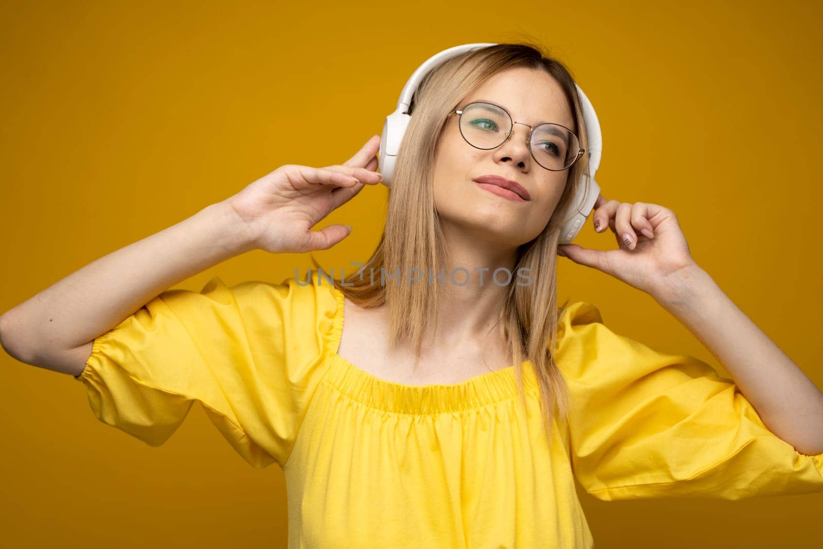 Beautiful attractive young blond woman wearing yellow t-shirt and glasses in white headphones listening music, dancing and laughing on blue background in studio. Relaxing and enjoying. Lifestyle