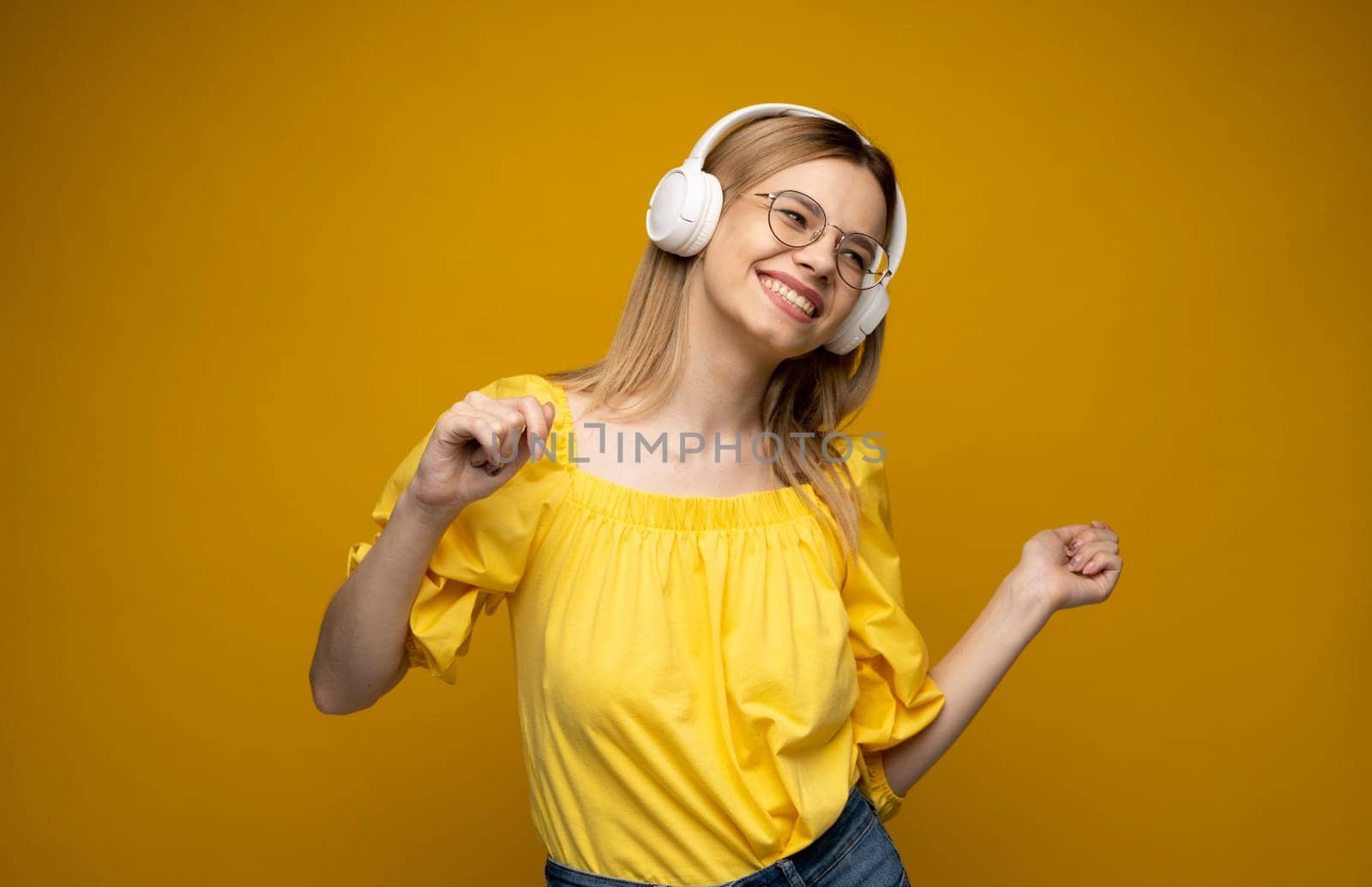 Cheerful young pretty girl smiling while listening music in headphones and dancing on yellow background