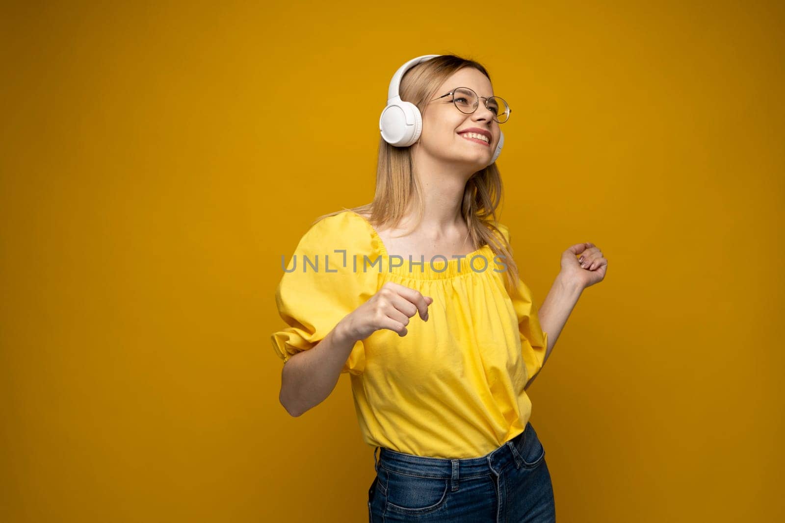 Cheerful young pretty girl smiling while listening music in headphones and dancing on yellow background