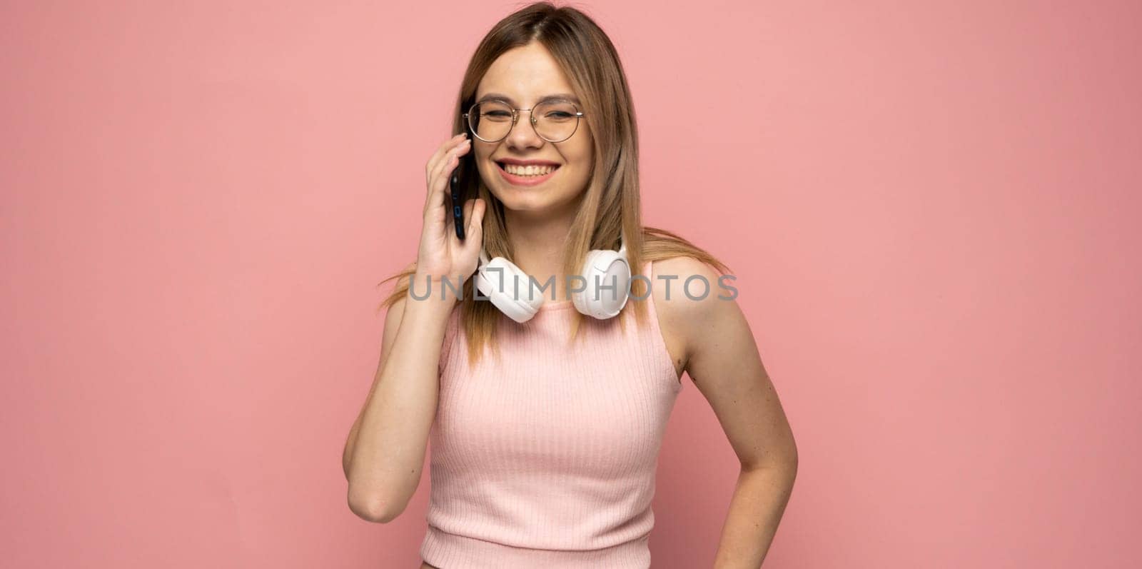 Portrait of a cute happy girl in yellow shirt and glasses with a headphones on a neck talking on mobile phone and laughing