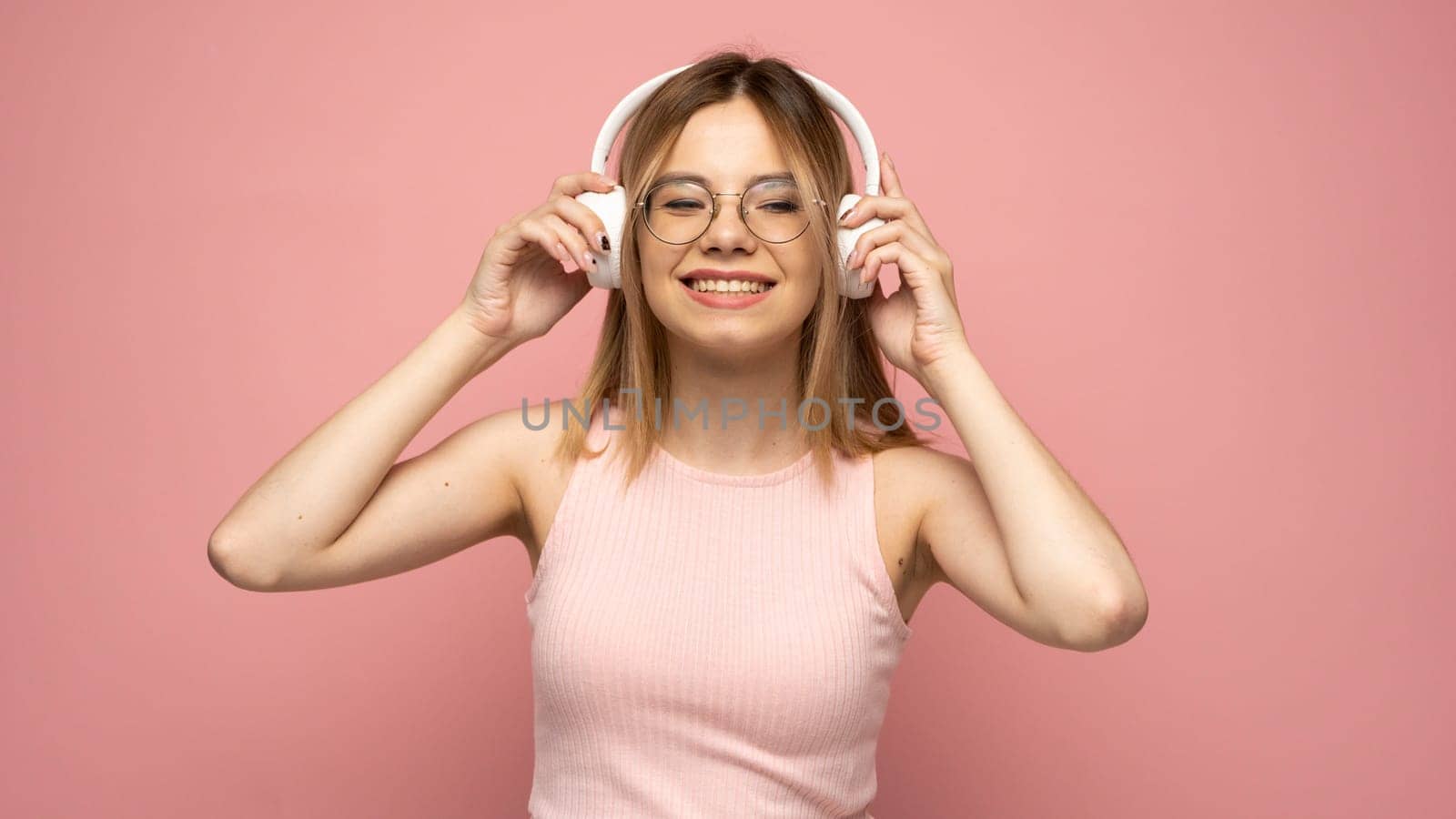 Beautiful attractive young blond woman wearing pink t-shirt and glasses in white headphones listening music and smiling on pink background in studio. Relaxing and enjoying. Lifestyle. by vovsht