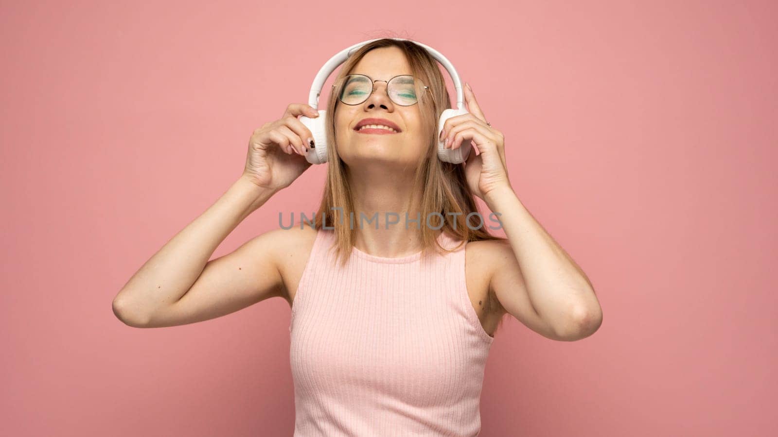 Beautiful attractive young blond woman wearing pink t-shirt and glasses in white headphones listening music and smiling on pink background in studio. Relaxing and enjoying. Lifestyle