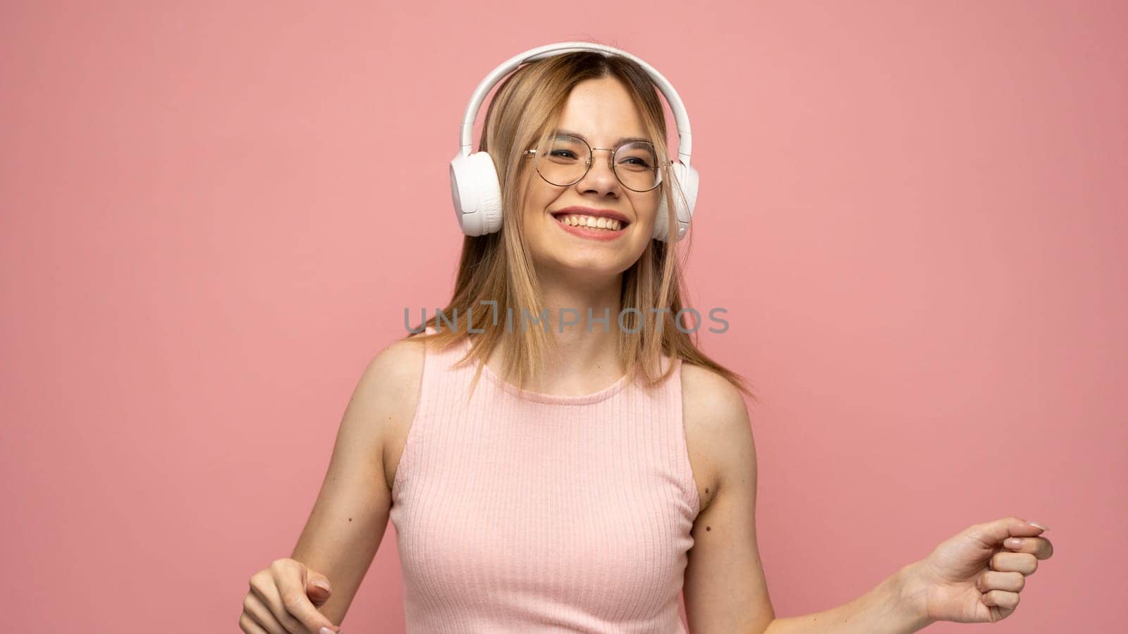 Pretty girl in a pink t-shirt and glasses listening music with her headphones and dancing on pink background