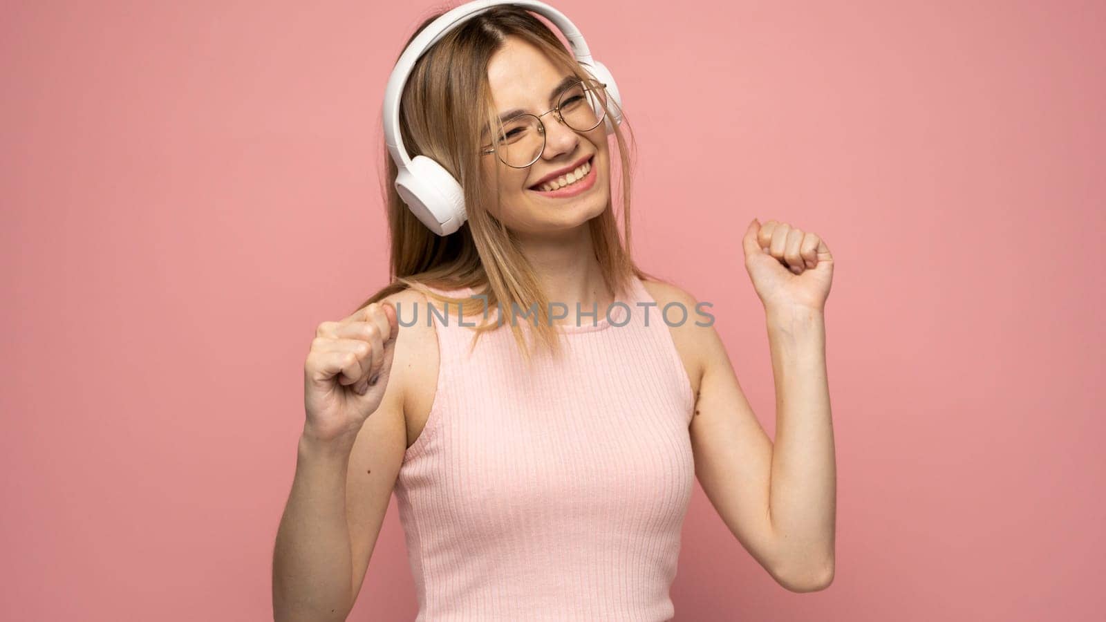 Blonde woman in a glasses and pink t-shirt listening music with a headphones. Teenager girl dancing against pink background
