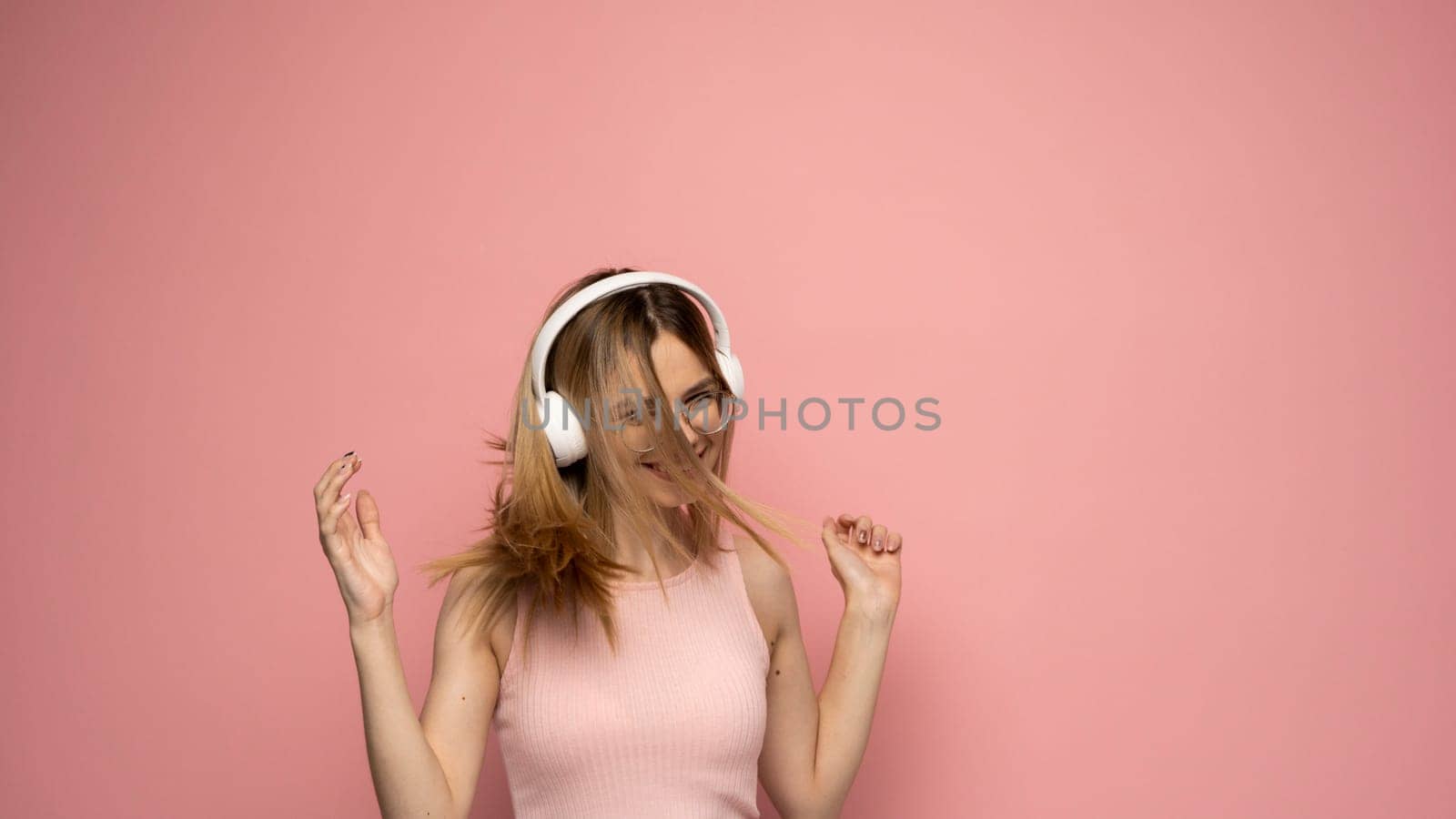 Beautiful attractive young blond woman wearing pink t-shirt and glasses in white headphones listening music and dancing on pink background in studio. Relaxing and enjoying. Lifestyle
