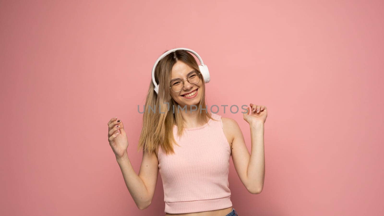 Beautiful attractive young blond woman wearing pink t-shirt and glasses in white headphones listening music and dancing on pink background in studio. Relaxing and enjoying. Lifestyle. by vovsht