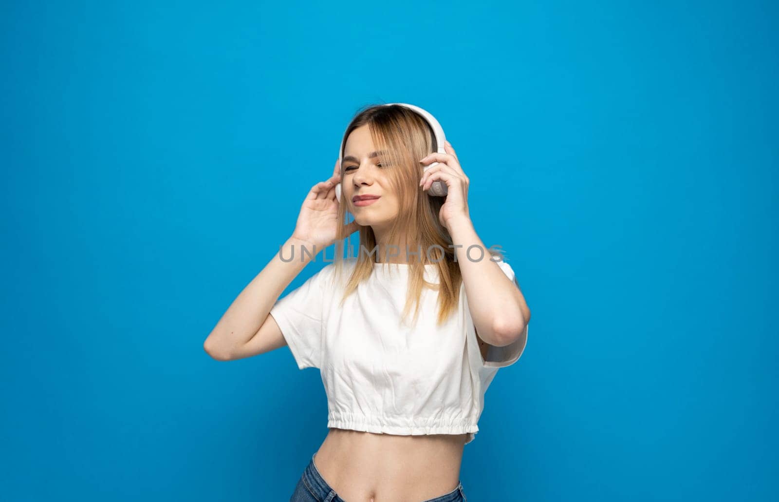 Cheerful young blonde woman girl in white clothes listening music with headphones, mobile phone isolated on blue background studio. People lifestyle concept. by vovsht