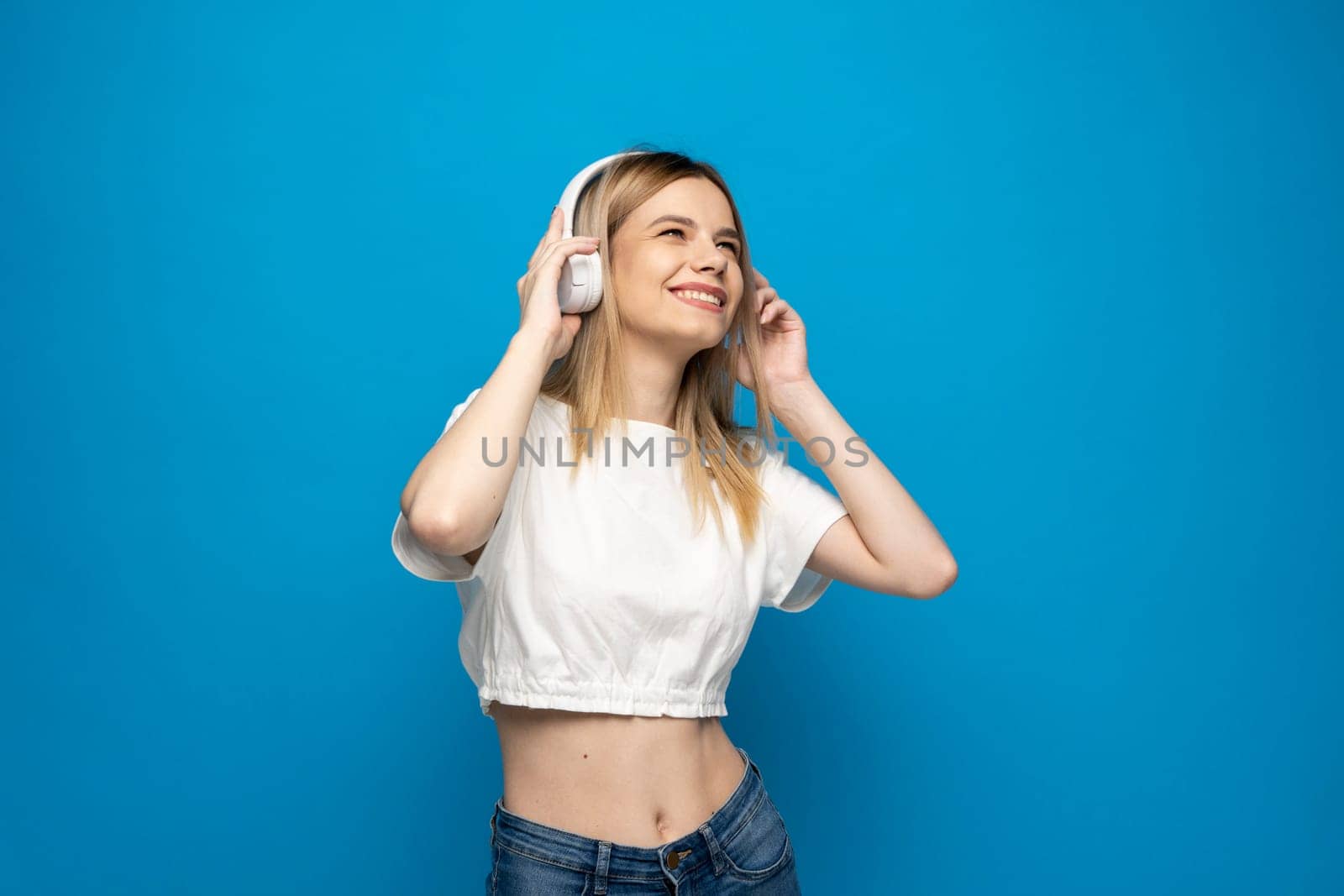 Beautiful attractive young blond woman wearing white t-shirt and glasses in white headphones listening music and dancing on blue background in studio. Relaxing and enjoying. Lifestyle. by vovsht