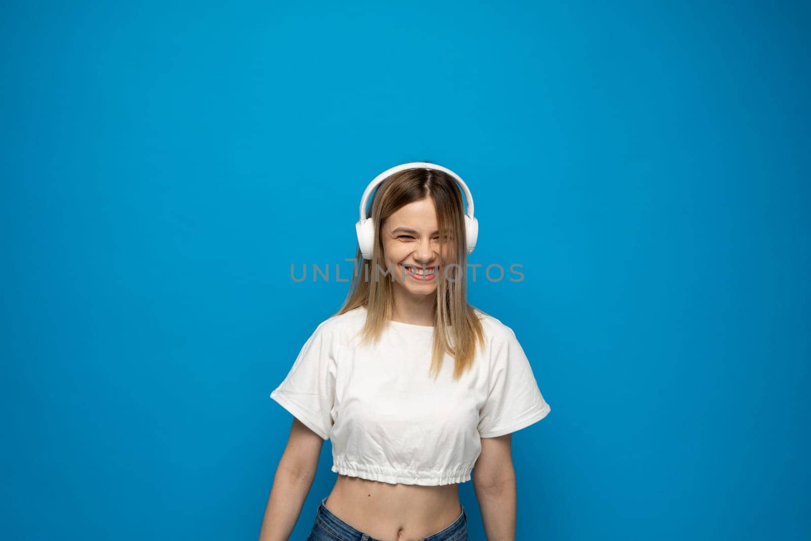 Smiling attractive young blond woman wearing white t-shirt and glasses in white headphones listening music and dancing on blue background in studio. Relaxing and enjoying. Lifestyle. by vovsht