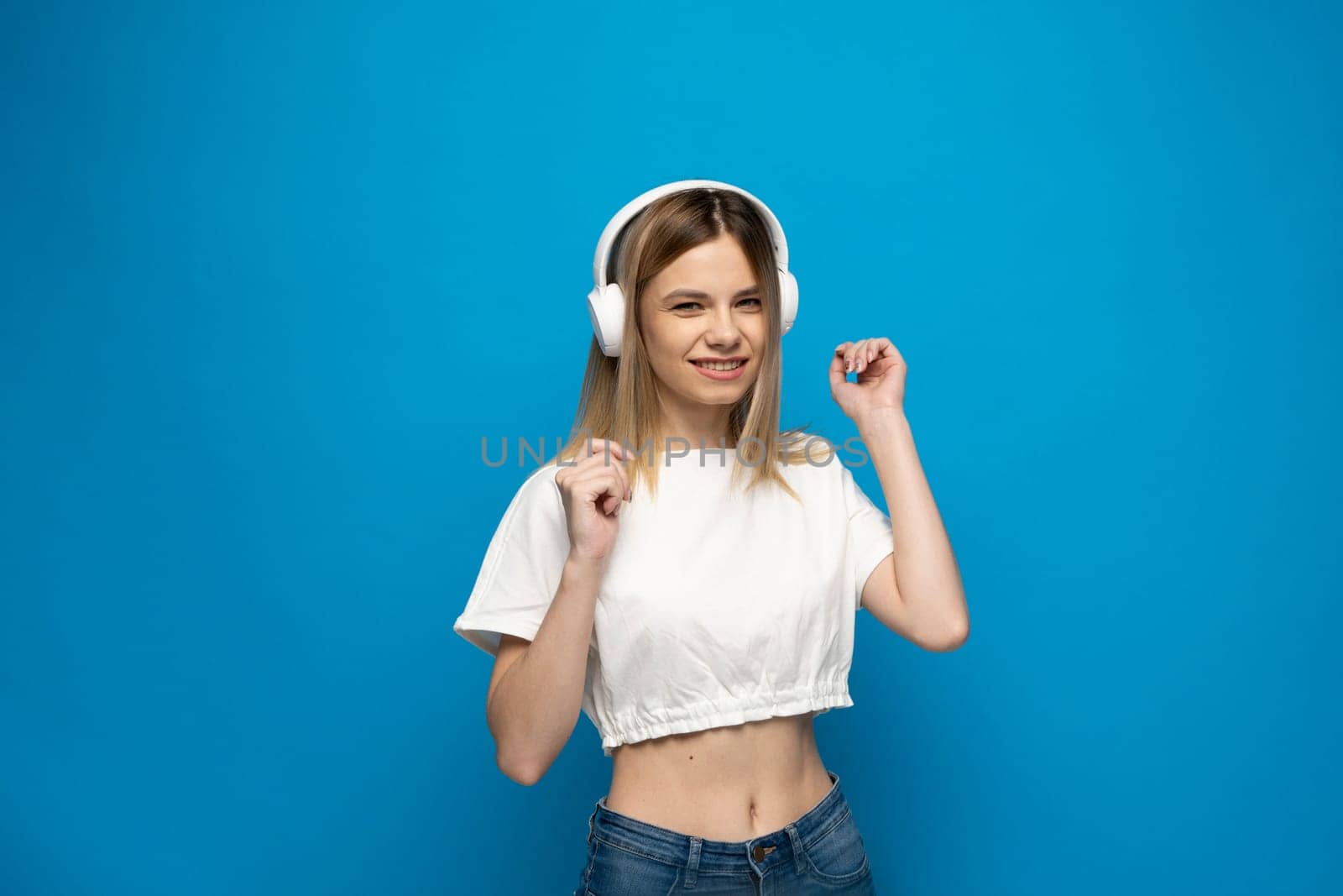 Cheerful young pretty girl smiling while listening music in headphones and dancing on blue background