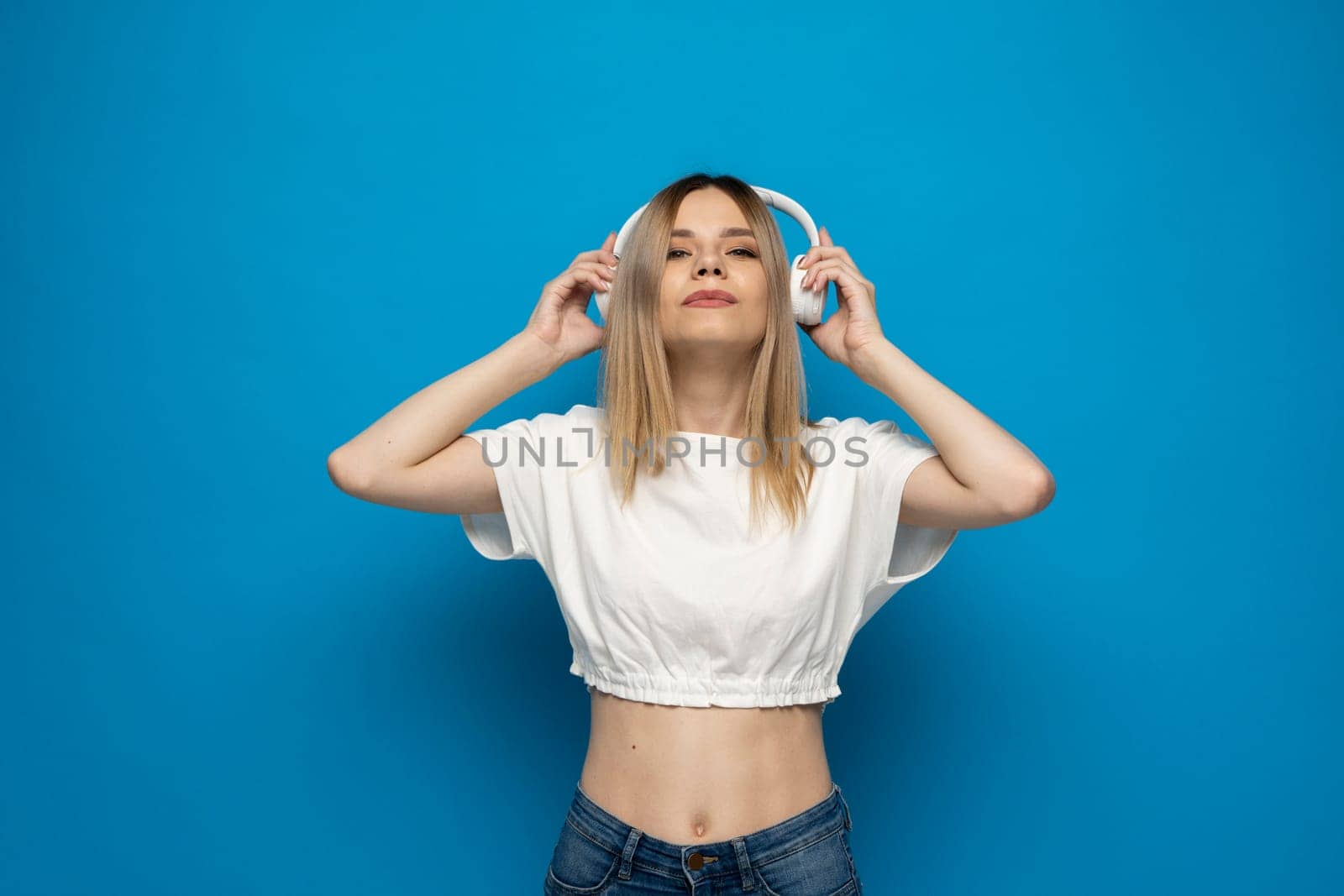 Beautiful attractive young blond woman wearing white t-shirt and glasses in white headphones listening music and smiling on blue background in studio. Relaxing and enjoying. Lifestyle. by vovsht