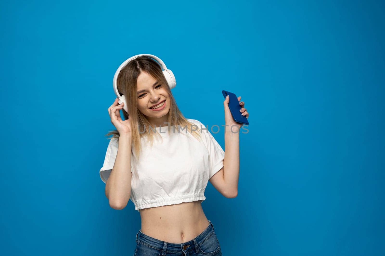 Beautiful attractive young blond woman wearing white t-shirt and glasses in white headphones listening music from smartphone, dancing and laughing on blue background in studio. Lifestyle. by vovsht