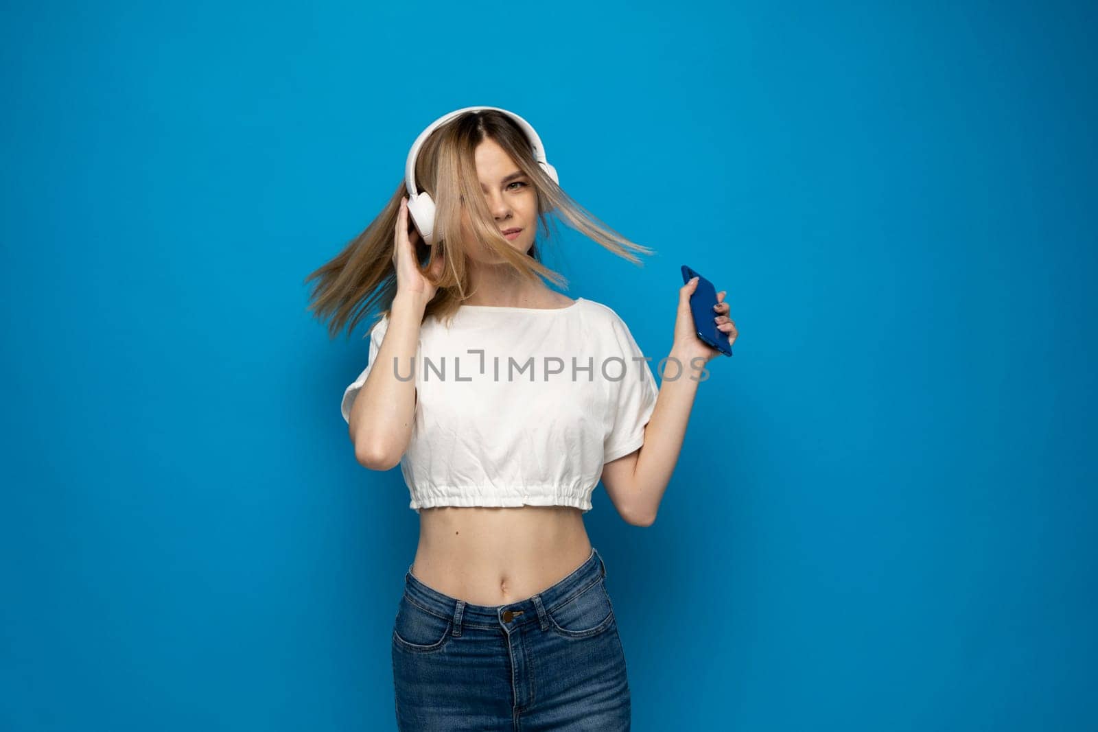 Beautiful young blonde woman with headphones and mobile device listening to music and smiling and dancing, isolated on blue background. by vovsht