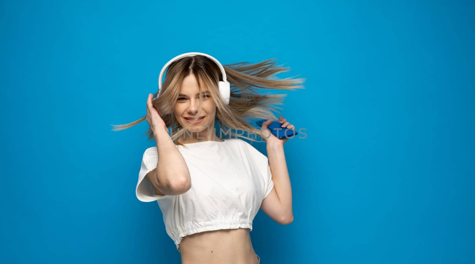 Beautiful attractive young blond woman wearing white t-shirt and glasses in white headphones listening music from smartphone, dancing and laughing on blue background in studio. Lifestyle. by vovsht