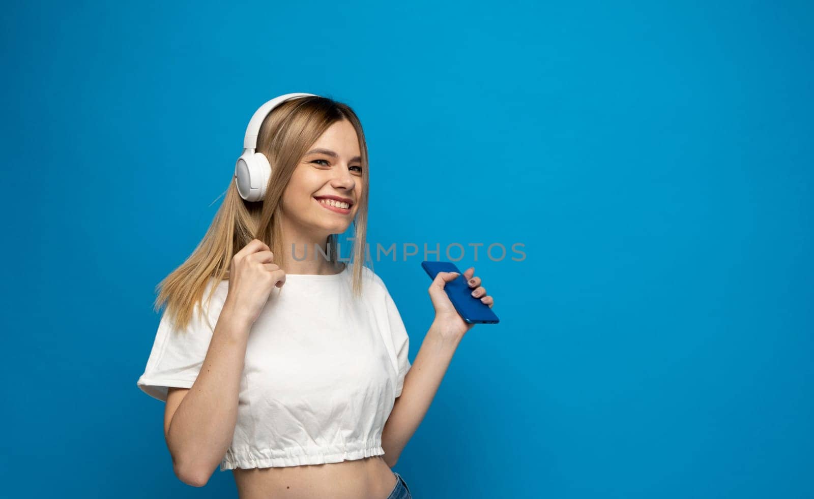 Beautiful attractive young blond woman wearing white t-shirt and glasses in white headphones listening music from smartphone, dancing and laughing on blue background in studio. Lifestyle
