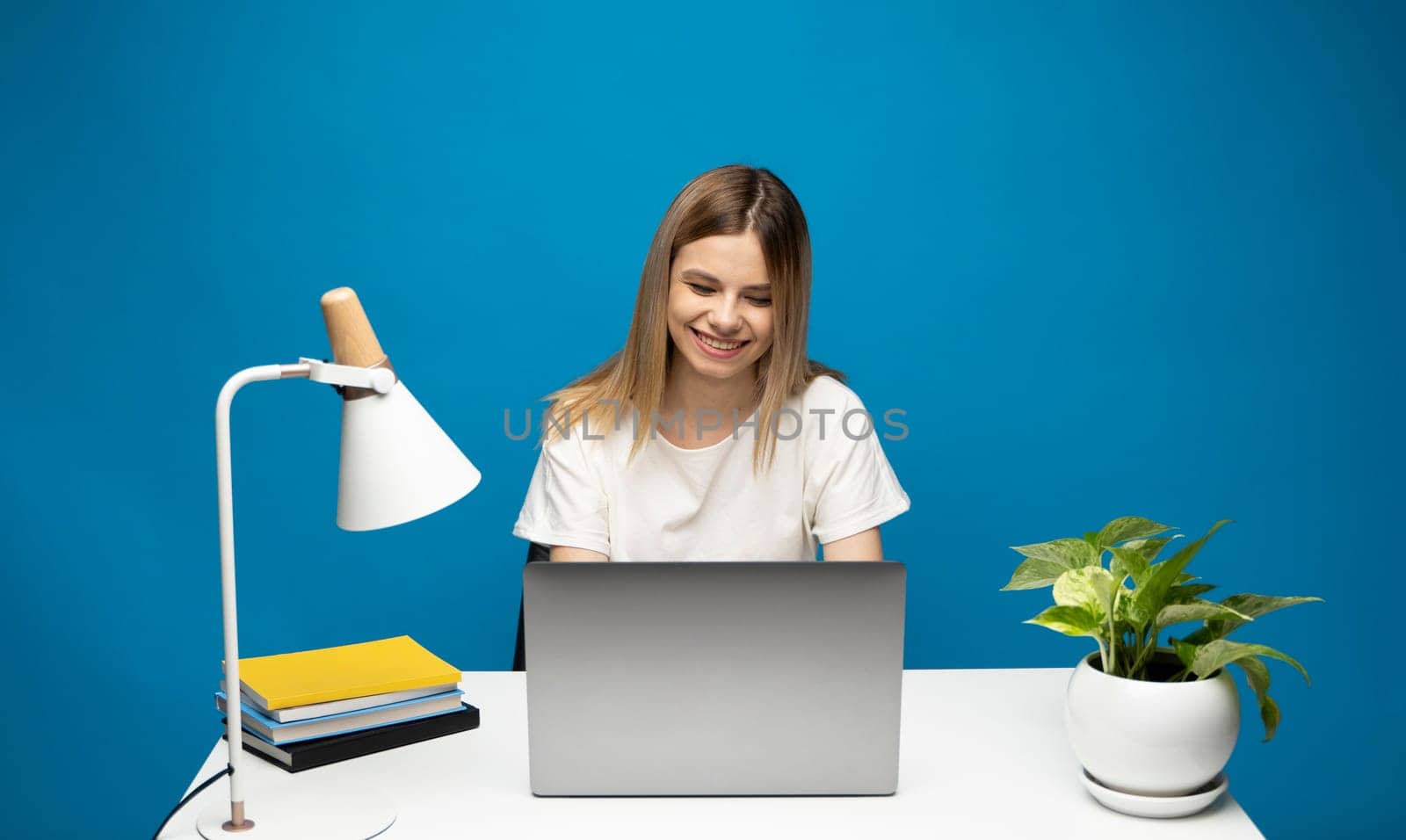 Excited young woman working with a grey laptop computer, notebook while sitting at the table with grey laptop. Smiling business woman or student received a good news isolated on a blue background