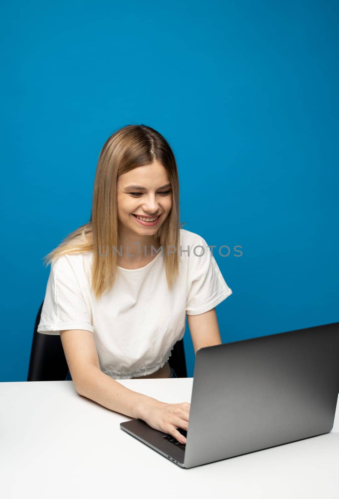 Portrait of a pretty young woman studying while sitting at the table with grey laptop computer, notebook. Smiling business woman working with a laptop isolated on a blue background. by vovsht