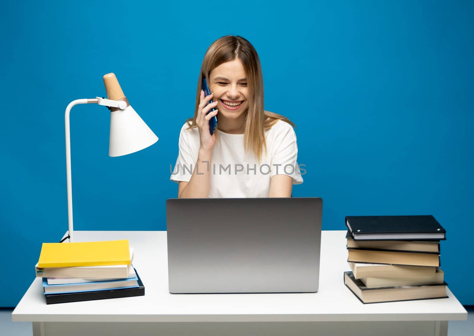 Young attractive business woman with a blond hair sitting at the table and working on a laptop and talking with a client on the phone. Young cheerful student girl talking with a smartphone. Studying. by vovsht