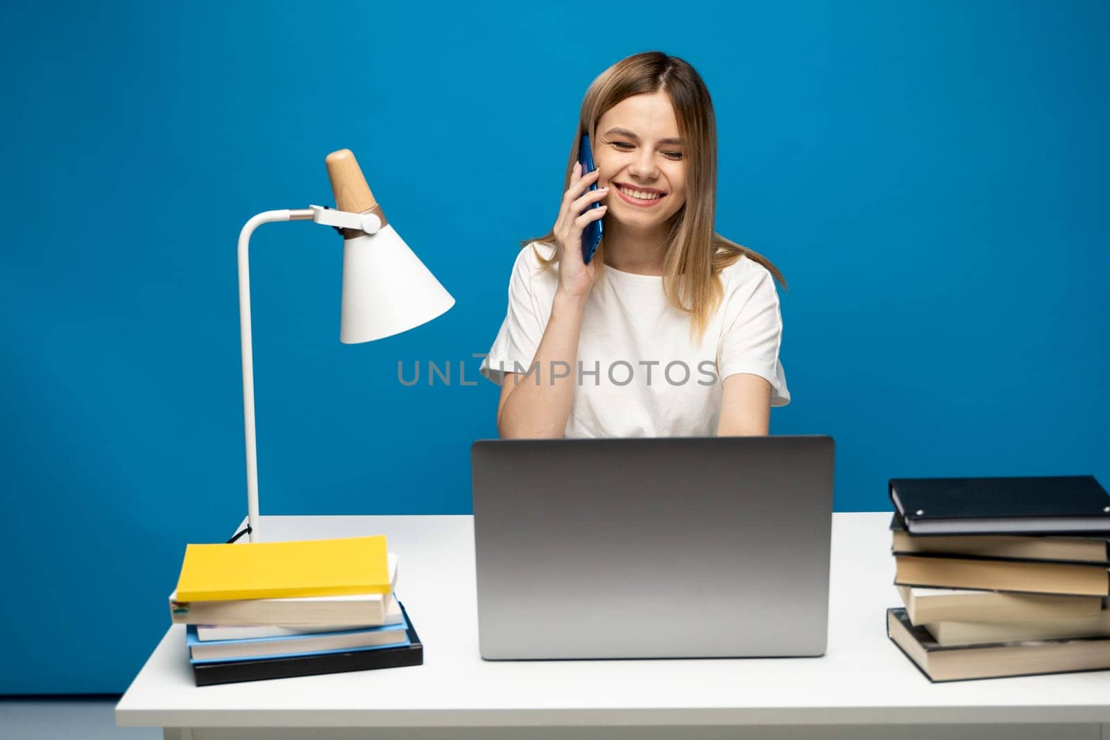 Young attractive business woman with a blond hair sitting at the table and working on a laptop and talking with a client on the phone. Young cheerful student girl talking with a smartphone. Studying