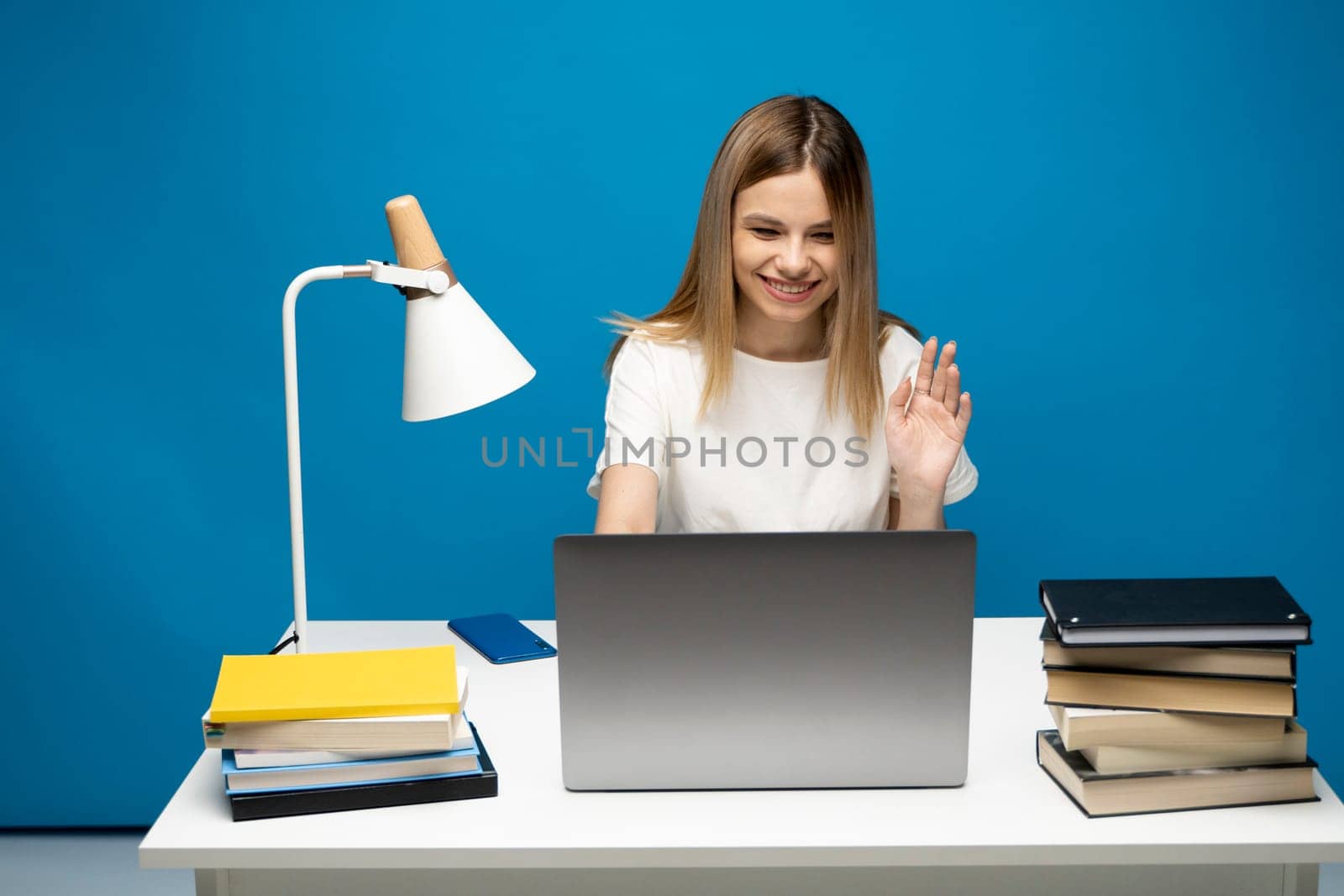 Front view young female freelancer sitting at studio before laptop screen waving hand to web camera. Student starting distant lesson, lecture or training course online. by vovsht