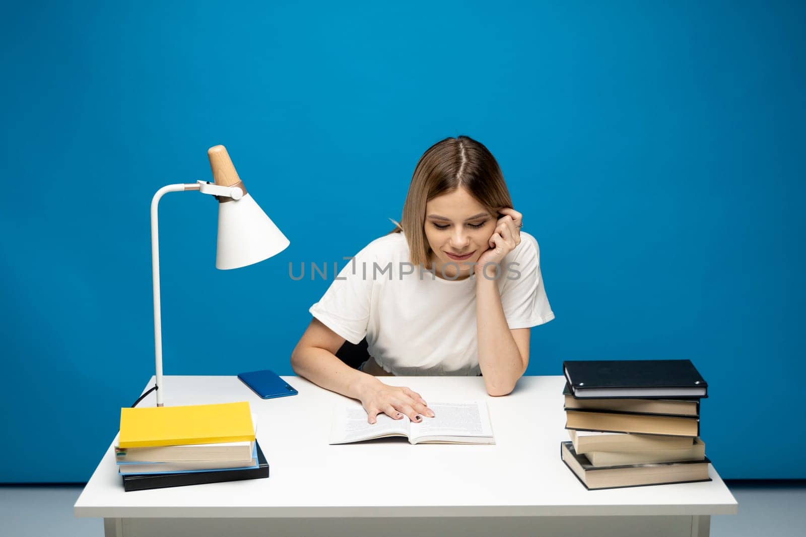 Young student woman in white casual clothes reading the book in library of university or college. Sitting and reading on blue background. Studying