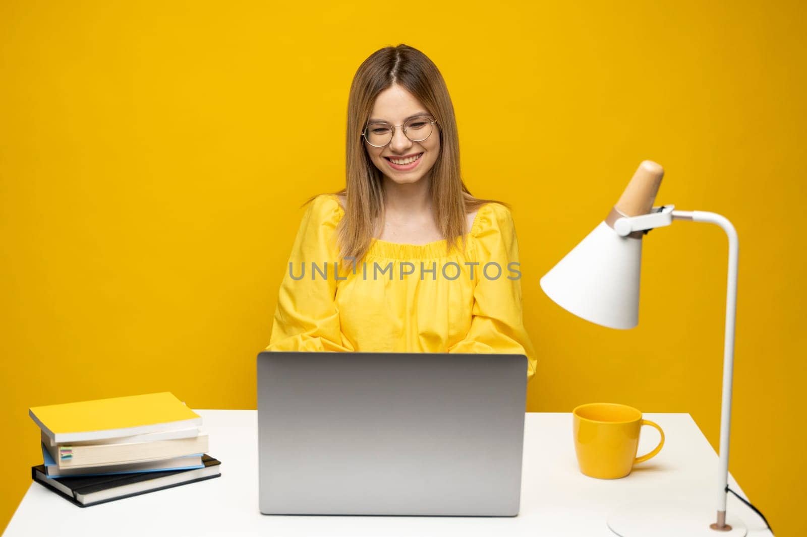 Excited young woman working with a grey laptop computer, notebook while sitting at the table with grey laptop. Smiling business woman or student received a good news isolated on a yellow background