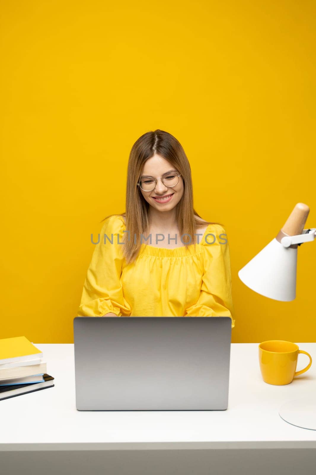 Portrait of Smiling pretty young woman studying while sitting at the table with grey laptop computer, notebook. Business woman working with a laptop isolated on a yellow background