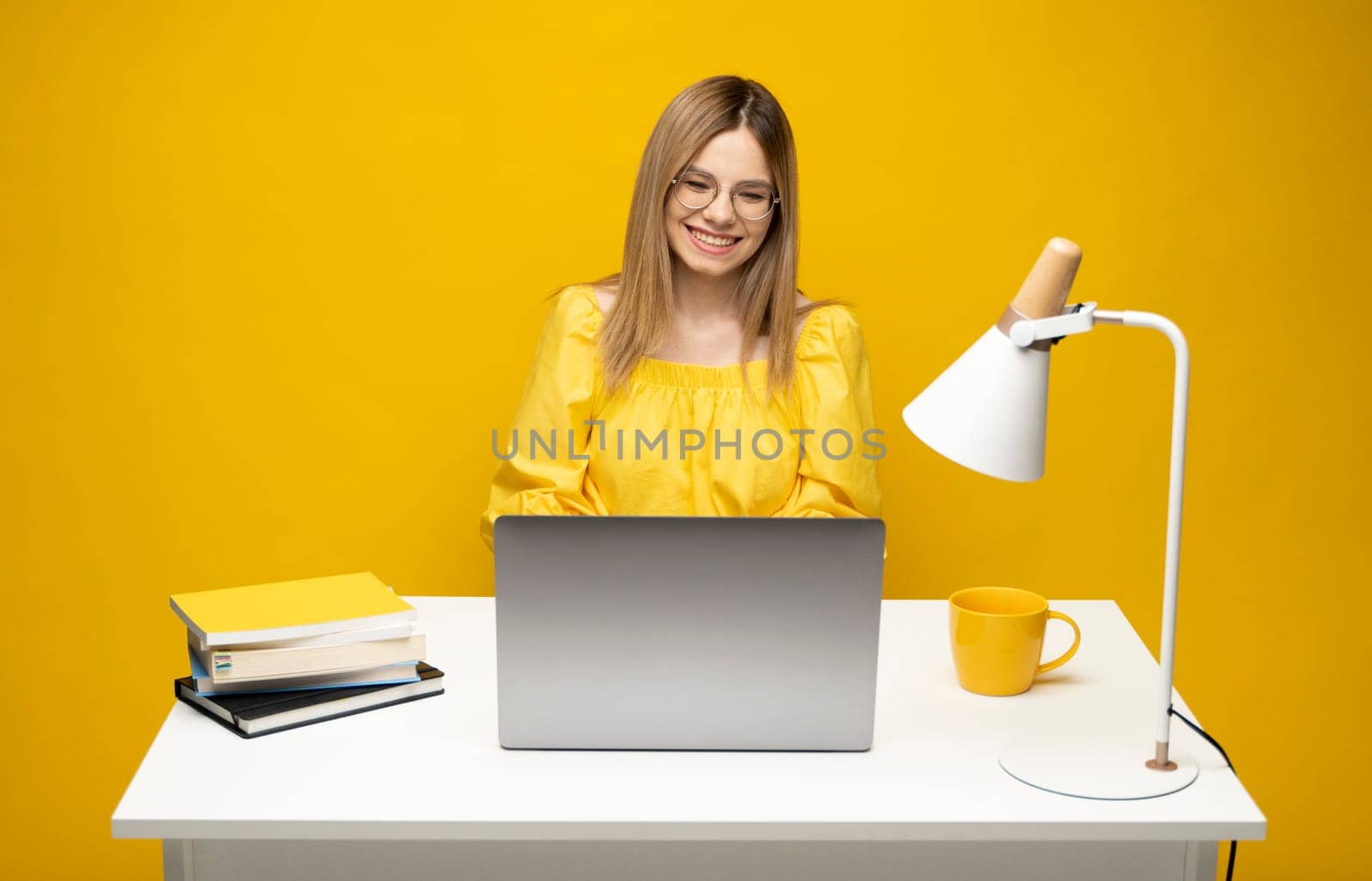 Portrait of a pretty young woman studying while sitting at the table with grey laptop computer, notebook. Smiling business woman working with a laptop isolated on a yellow background