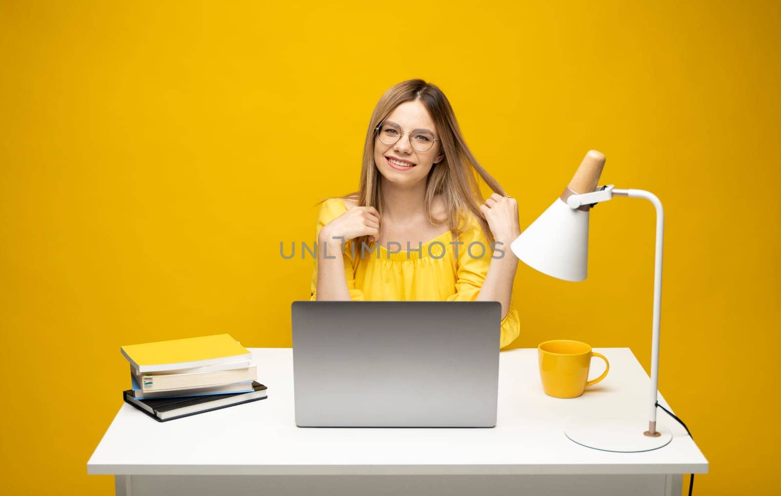 Portrait of Smiling pretty young woman studying while sitting at the table with grey laptop computer, notebook. Business woman working with a laptop isolated on a yellow background