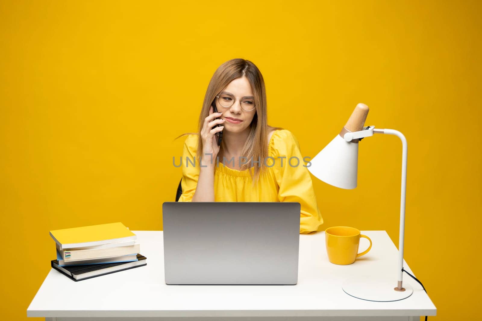 Young attractive business woman with a blond hair sitting at the table and working on a laptop and talking with a client on the phone. Young cheerful student girl talking with a smartphone. Studying