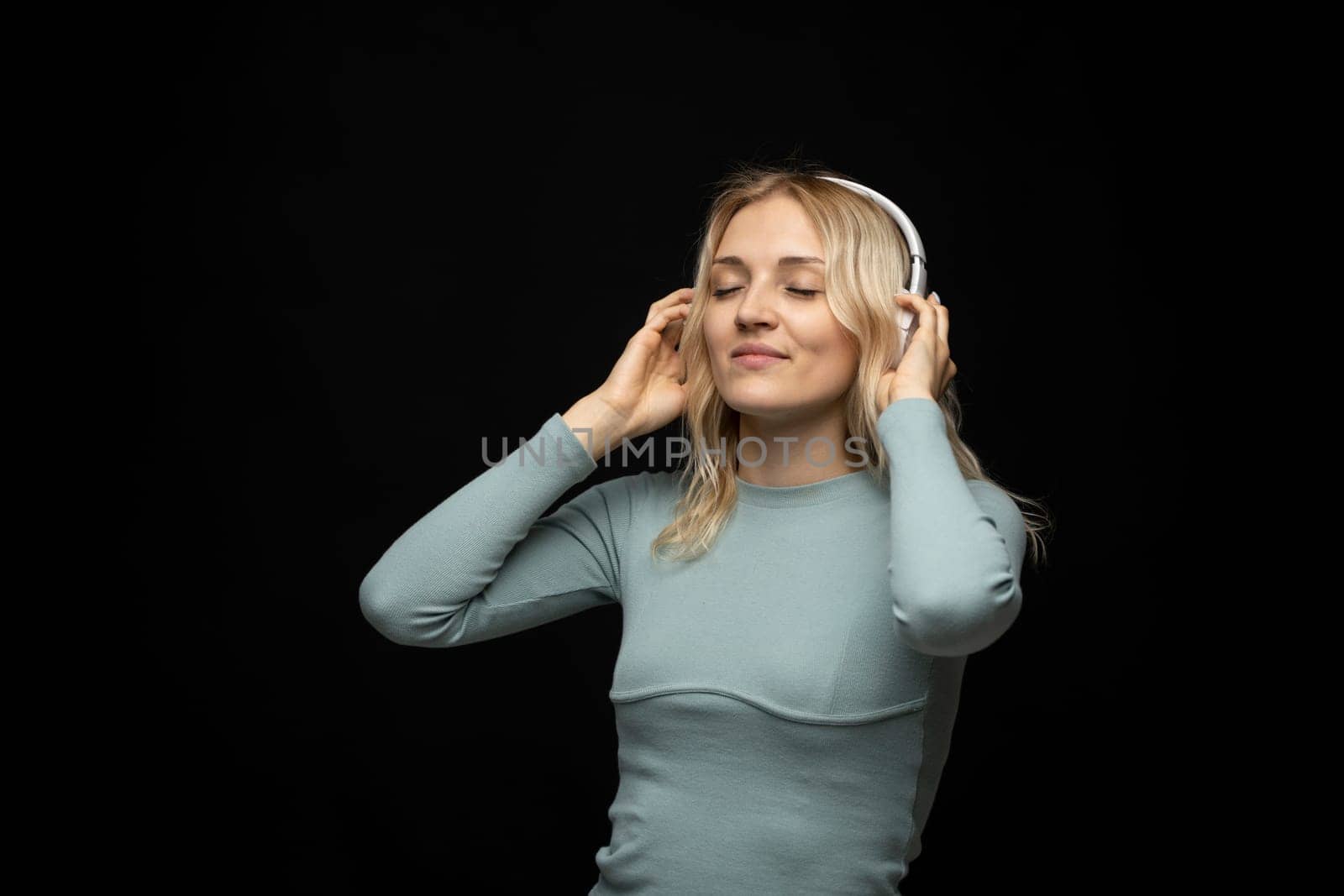 Beautiful attractive young blond woman wearing blue t-shirt and glasses in white headphones listening music and smiling on black background in studio. Relaxing and enjoying. Lifestyle