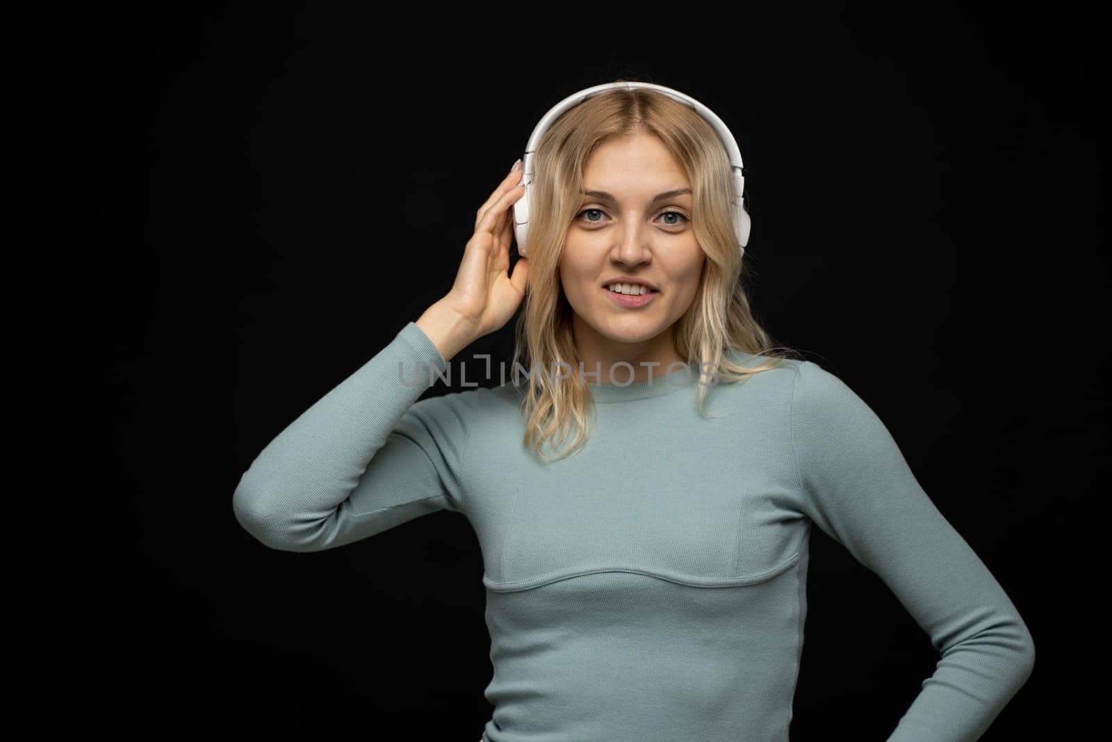 Beautiful attractive young blond woman wearing blue t-shirt and glasses in white headphones listening music and smiling on black background in studio. Relaxing and enjoying. Lifestyle. by vovsht