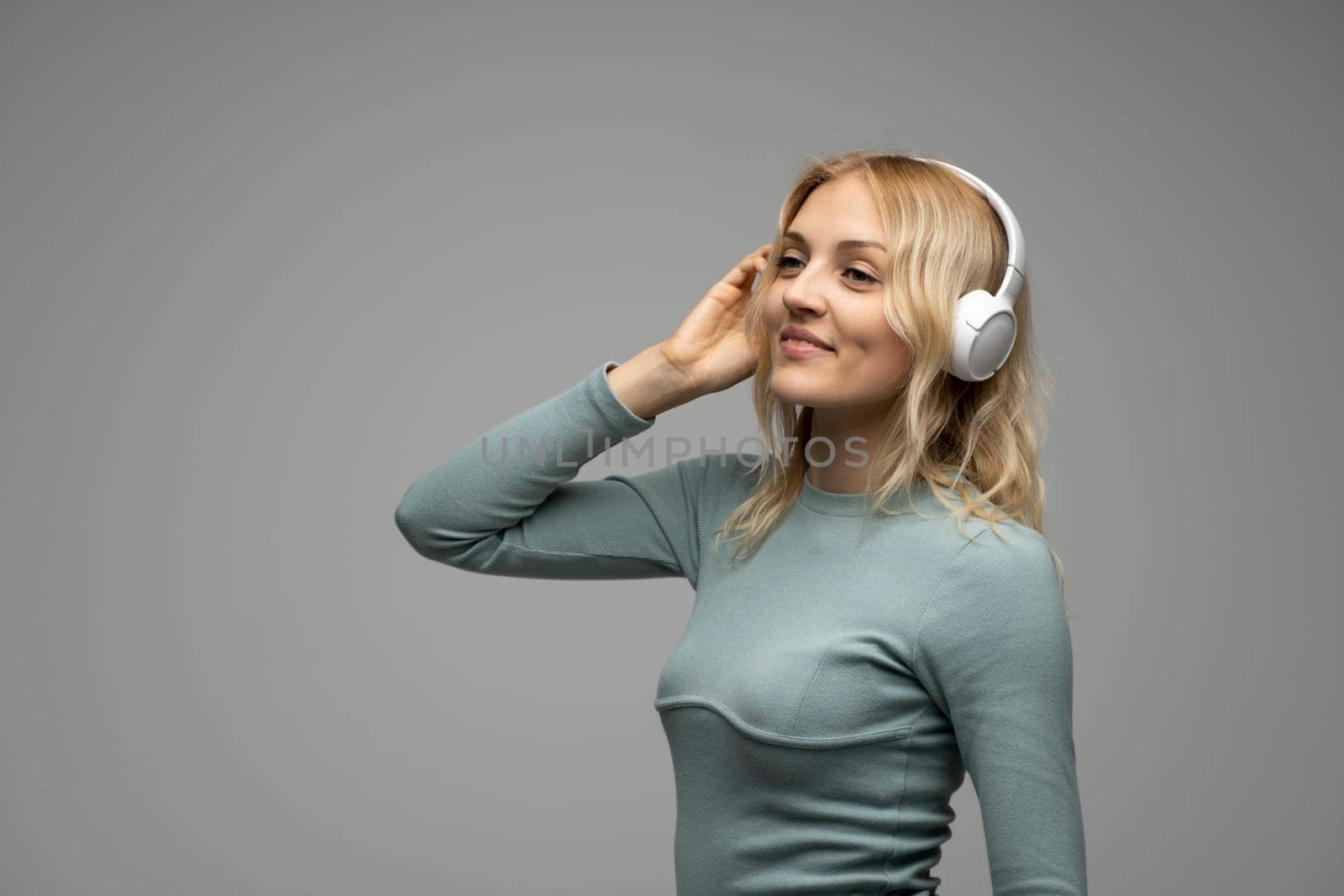 Blonde girl with white headphones listening to music on grey background in studio