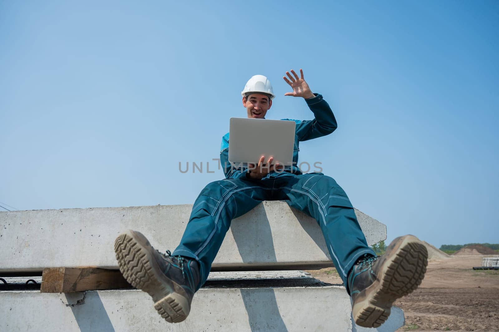 Caucasian male builder in hardhat sits on floor slabs and uses laptop at construction site