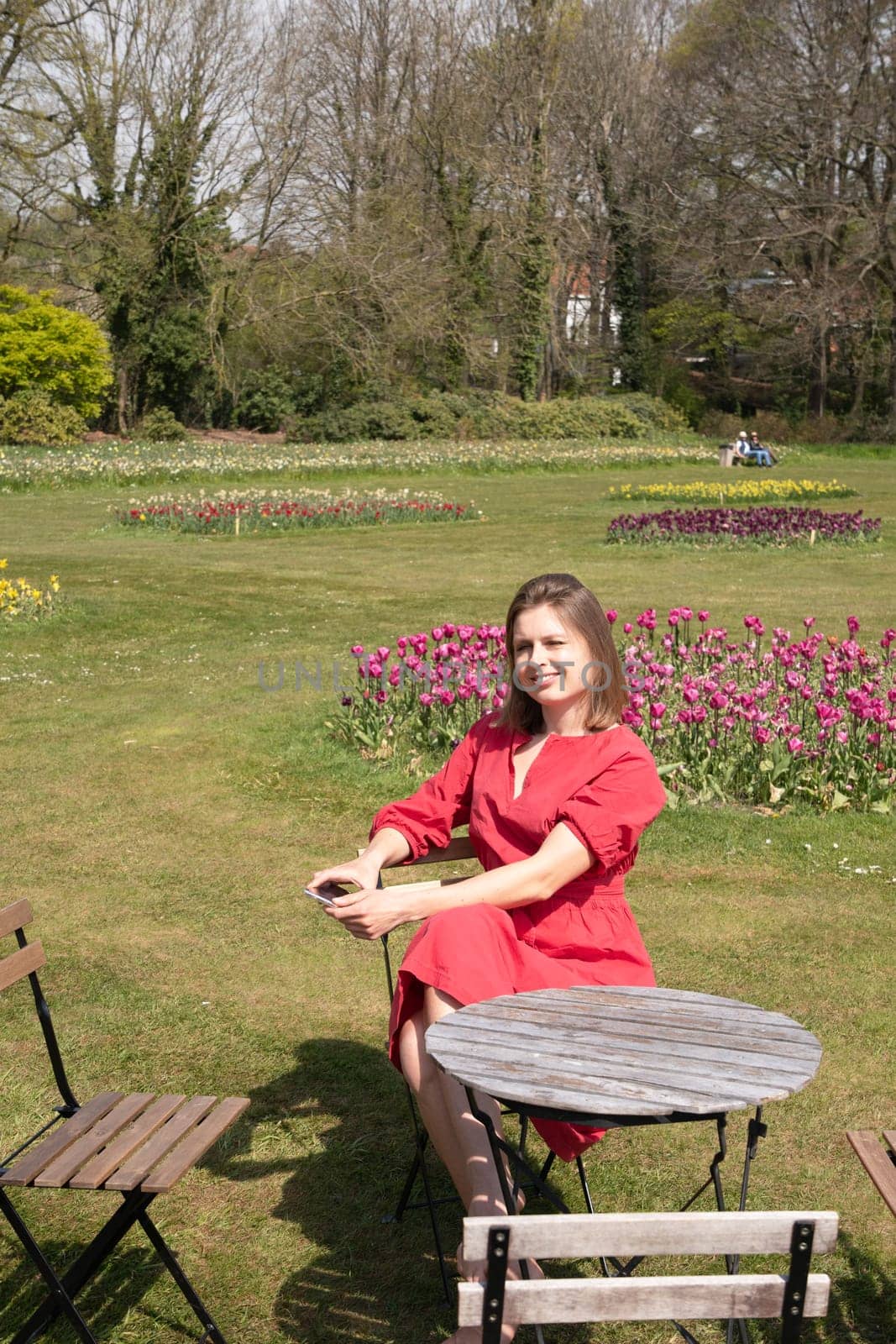 A young beautiful girl in a red dress in a spring park plays with her hair, flirts and takes a selfie on a mobile phone, tulips bloom and people walk in the park, pamper themselves.High quality photo