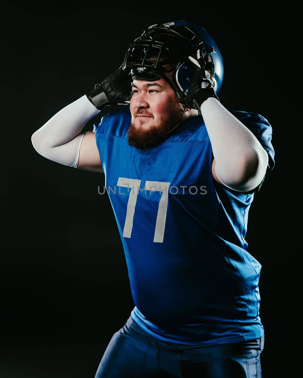 An Asian man with a red beard in a blue american football uniform puts on a helmet on a black background